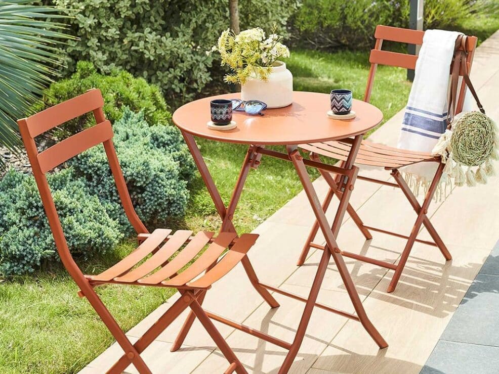 Conjunto de mesa y sillas plegables de jardín en un rincón exterior con plantas, de Fesmés